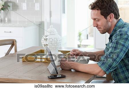 Stock Image - Mid adult man eating breakfast, whilst looking at digital tablet. Fotosearch
