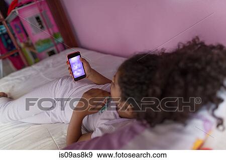 Over shoulder view of girl wearing pyjamas lying on bed looking at smartphone View Large Photo Image Stock Photo - Over shoulder view of girl wearing pyjamas lying on bed looking at smartphone. Fotosearch