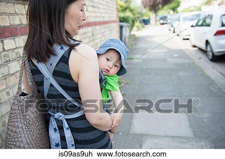 Stock Image - Portrait of baby boy being carried by mother in baby sling along street. Fotosearch