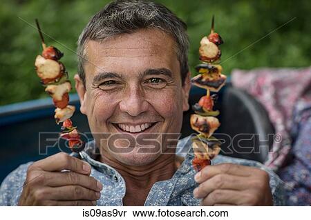 Stock Photography - Portrait of mature man holding barbecued kebab sticks. Fotosearch