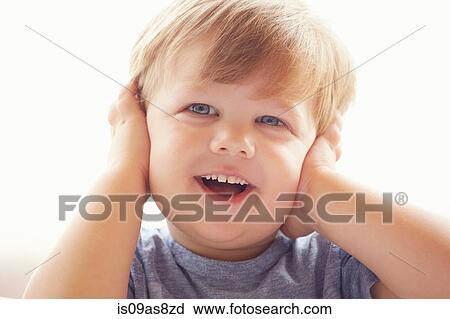 Stock Image - Portrait of young boy covering ears with hands, looking away smiling. Fotosearch