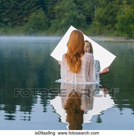 Stock Image - Rear view of young woman standing in lake holding mirror looking at reflection. Fotosearch