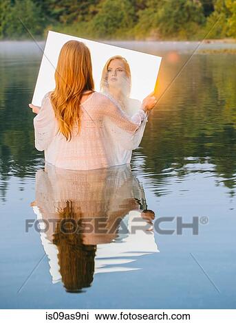 Stock Image - Rear view of young woman standing in lake holding mirror looking at reflection, lens flare. Fotosearch