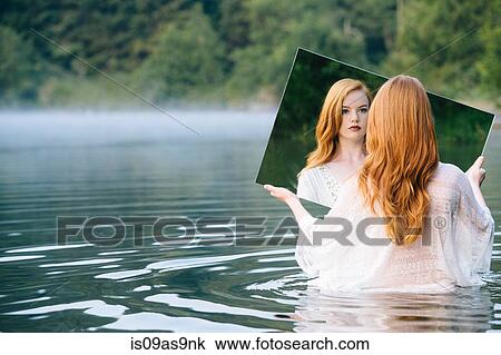 Stock Image - Rear view of young woman standing in lake holding mirror looking at reflection. Fotosearch
