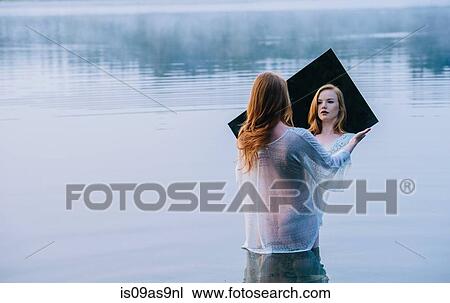 Stock Image - Rear view of young woman standing in lake holding mirror looking at reflection. Fotosearch