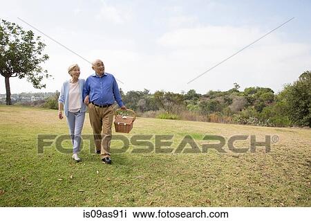 Senior couple walking arm in arm in park, carrying picnic basket View Large Photo Image Stock Image - Senior couple walking arm in arm in park, carrying picnic basket. Fotosearch