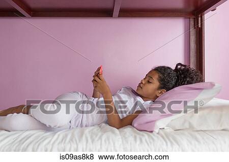 Stock Photo - Side view of girl wearing pyjamas lying on bed looking at smartphone. Fotosearch