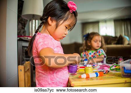 Stock Image - Two young girls sitting at table, making art, using paint. Fotosearch