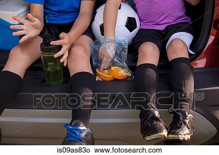 Waist down of boy and younger sister sitting in car boot eating oranges on football practice break View Large Photo Image Picture - Waist down of boy and younger sister sitting in car boot eating oranges on football practice break. Fotosearch