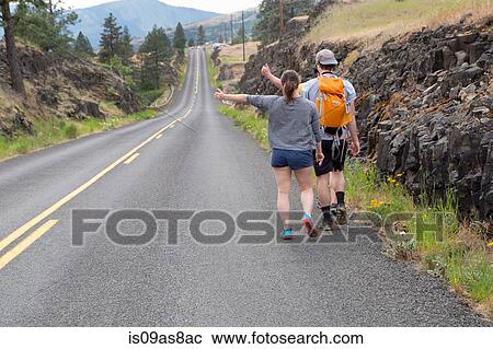 Stock Image - Young couple hiking along road, trying to thumb lift, rear view. Fotosearch