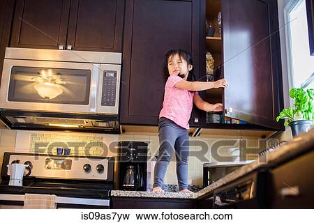 Stock Photo - Young girl standing on kitchen work surface looking for treats in cupboard. Fotosearch