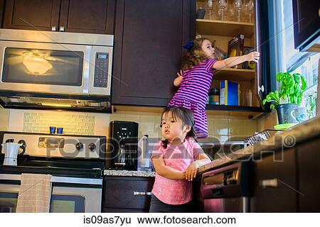 Stock Photograph - Young girl standing on kitchen work surface, looking for treats in cupboard. Fotosearch