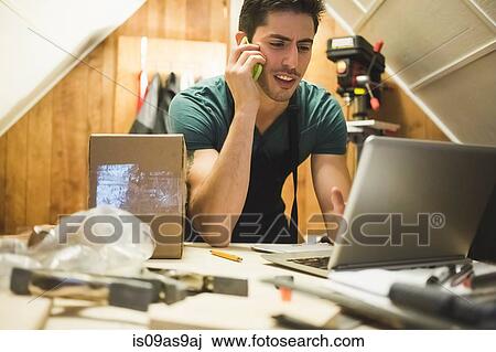 Young man in workshop sitting at desk talking on telephone looking at laptop View Large Photo Image Stock Image - Young man in workshop sitting at desk talking on telephone looking at laptop. Fotosearch