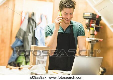 Young man in workshop standing at desk talking on telephone looking at laptop View Large Photo Image Stock Image - Young man in workshop standing at desk talking on telephone looking at laptop. Fotosearch