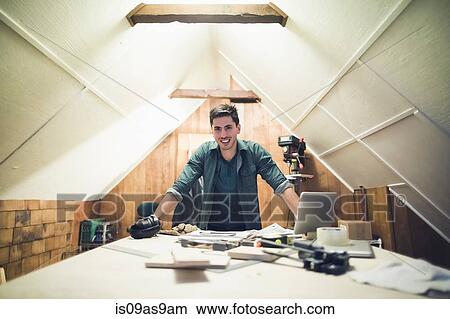 Picture - Young man leaning against desk in workshop looking at camera smiling. Fotosearch