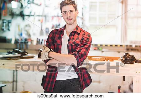 Young man leaning against workbench wearing protective gloves, arms folded looking at camera View Large Photo Image Stock Photograph - Young man leaning against workbench wearing protective gloves, arms folded looking at camera. Fotosearch