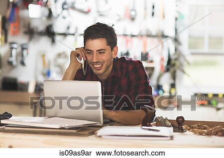 Young man sitting at desk in workshop talking on phone using laptop smiling View Large Photo Image Stock Photography - Young man sitting at desk in workshop talking on phone using laptop smiling. Fotosearch