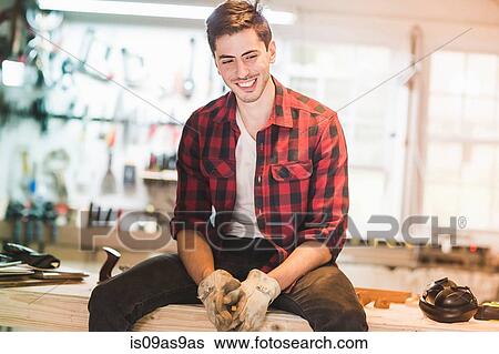 Stock Photograph - Young man sitting on workbench wearing protective gloves, looking away smiling. Fotosearch