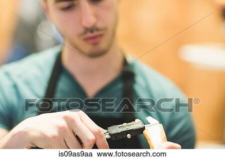 Stock Image - Young man wearing apron crafting wood object with tool, looking down. Fotosearch