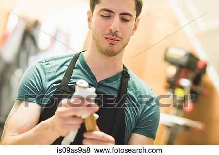 Stock Photo - Young man wearing apron crafting wood object, looking down. Fotosearch