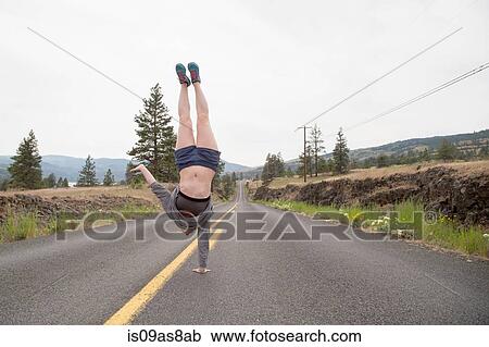 Stock Image - Young woman doing one handed handstand in road. Fotosearch