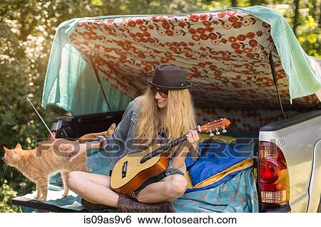 Stock Photograph - Young woman petting cat and playing ukulele whilst camping in pick up boot. Fotosearch