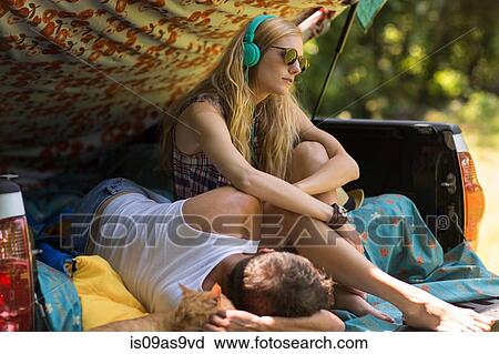 Stock Image - Young woman wearing headphones with boyfriend in pick up boot whilst camping. Fotosearch