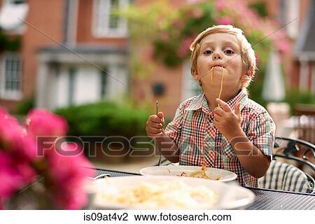Stock Image - Boy sitting at garden table sucking up spaghetti looking at camera. Fotosearch