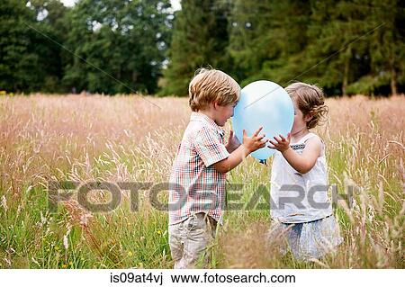 Stock Image - Brother and sister in tall grass face to face playing with balloon. Fotosearch