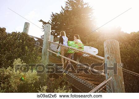 Stock Photograph - "Couple walking up beach steps, carrying surfboards". Fotosearch