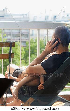 Stock Photo - Father using smartphone and cradling sleeping son on balcony. Fotosearch