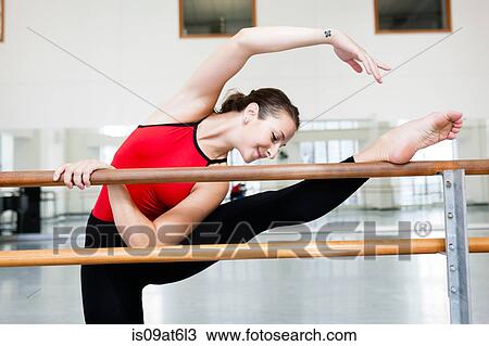 Stock Image - Front view of young woman in dance studio leg raised stretching on barre. Fotosearch