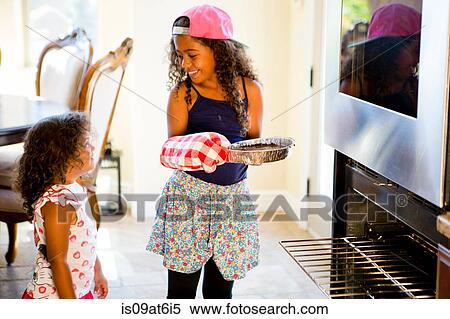 "Girl watching big sister insert cake tin into oven, face to face smiling" View Large Photo Image Stock Photography - "Girl watching big sister insert cake tin into oven, face to face smiling". Fotosearch