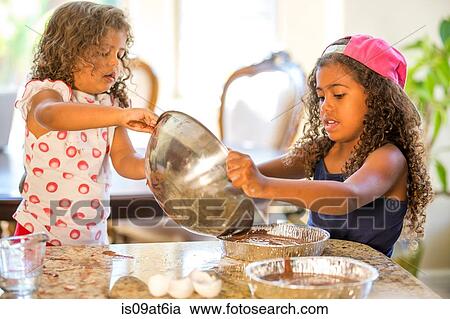 Stock Image - Girls pouring cake mix from mising bowl into cake tins. Fotosearch