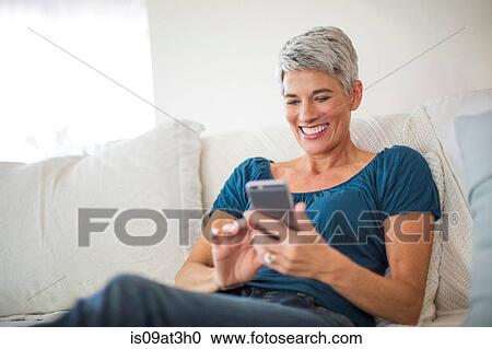 Stock Image - Happy mature woman reclining on sofa texting on smartphone. Fotosearch
