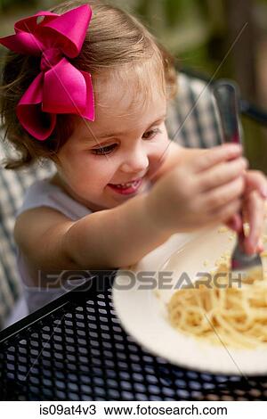 Stock Image - High angle vew of girl with pink hair bow sitting at garden eating spaghetti. Fotosearch