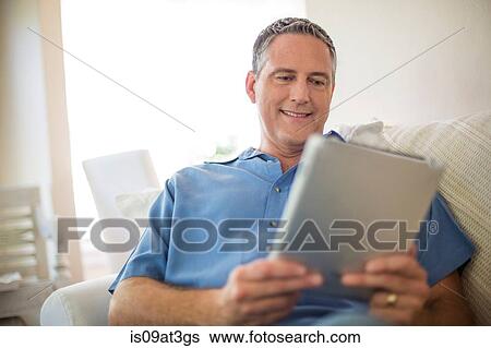 Stock Photograph - Mature man reading digital tablet on sofa. Fotosearch