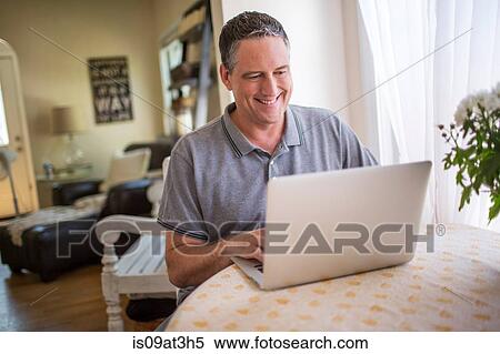Stock Photography - Mature man typing on laptop at kitchen table. Fotosearch