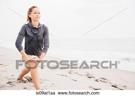 Stock Image - Mid adult woman doing stretches on beach. Fotosearch