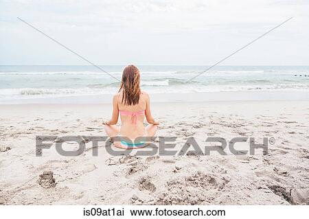Stock Image - "Mid adult woman meditating on beach, rear view". Fotosearch