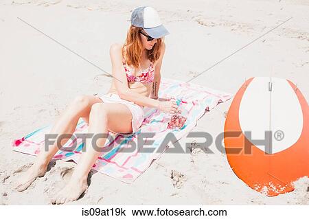 Stock Image - "Mid adult woman on beach, eating grapes, surfboard beside her". Fotosearch