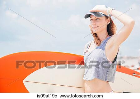 Stock Image - "Mid adult woman on beach, holding surfboard". Fotosearch