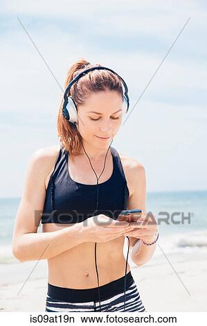 "Mid adult woman standing on beach, wearing headphones, looking at mp3 player" View Large Photo Image Picture - "Mid adult woman standing on beach, wearing headphones, looking at mp3 player". Fotosearch