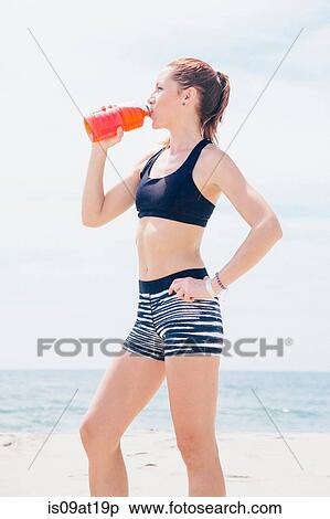 Stock Photography - "Mid adult woman standing on beach, drinking drink from plastic bottle". Fotosearch