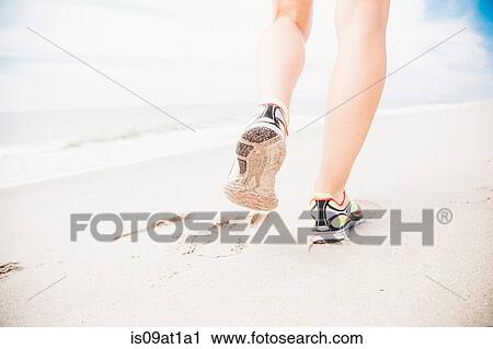 Stock Image - "Mid adult woman walking on beach, rear view, low section". Fotosearch
