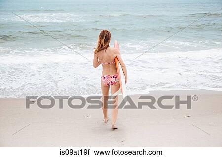 Stock Image - "Mid adult woman walking towards sea, holding surfboard, rear view". Fotosearch
