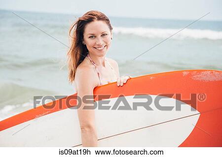 Stock Image - Portrait of mid adult woman holding surfboard. Fotosearch