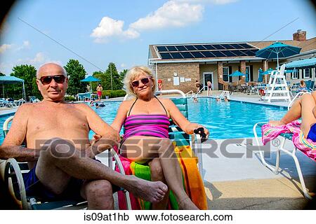 Stock Image - Portrait of smiling senior couple sitting poolside at outdoor swimming pool. Fotosearch