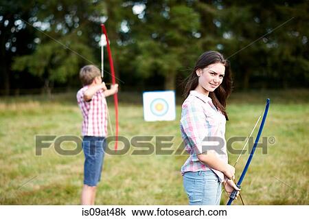 Stock Image - Portrait of teenage girl practicing archery with brother. Fotosearch