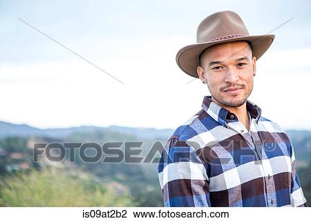 Stock Image - Portrait of young man wearing cowboy hat on rural hill. Fotosearch